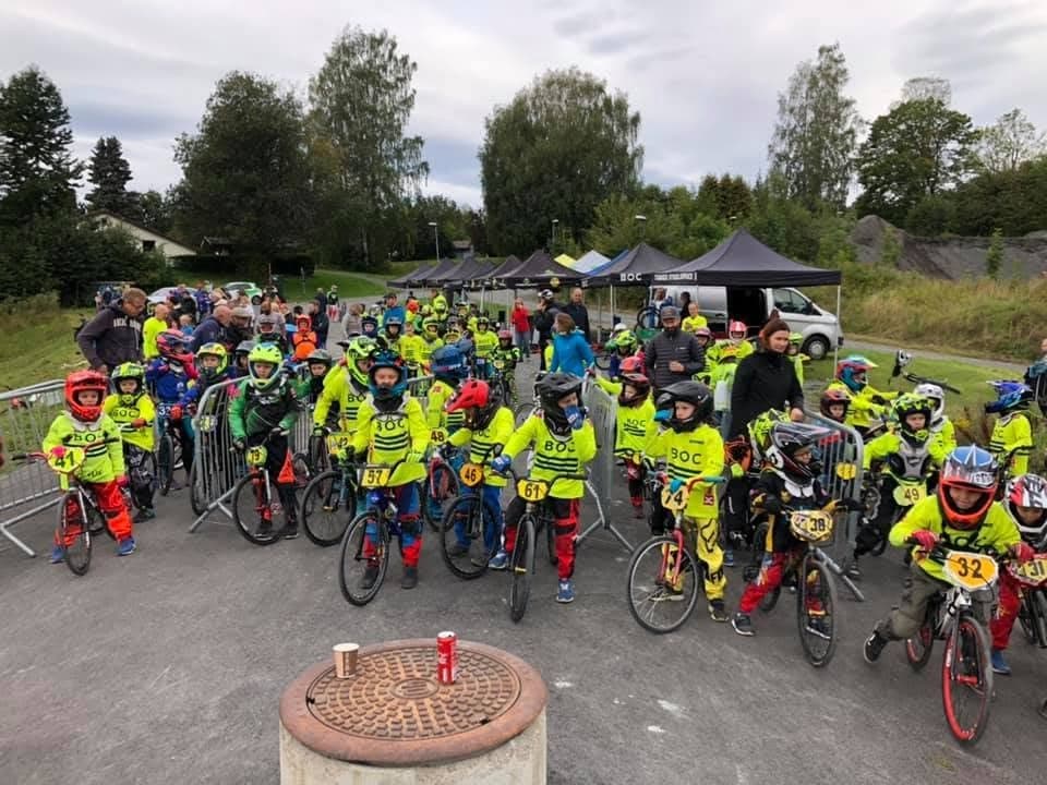 Mange barn i gul BOC-drakt og hjelm står klare på startstreken til et BMX-ritt, med foresatte og telt bak