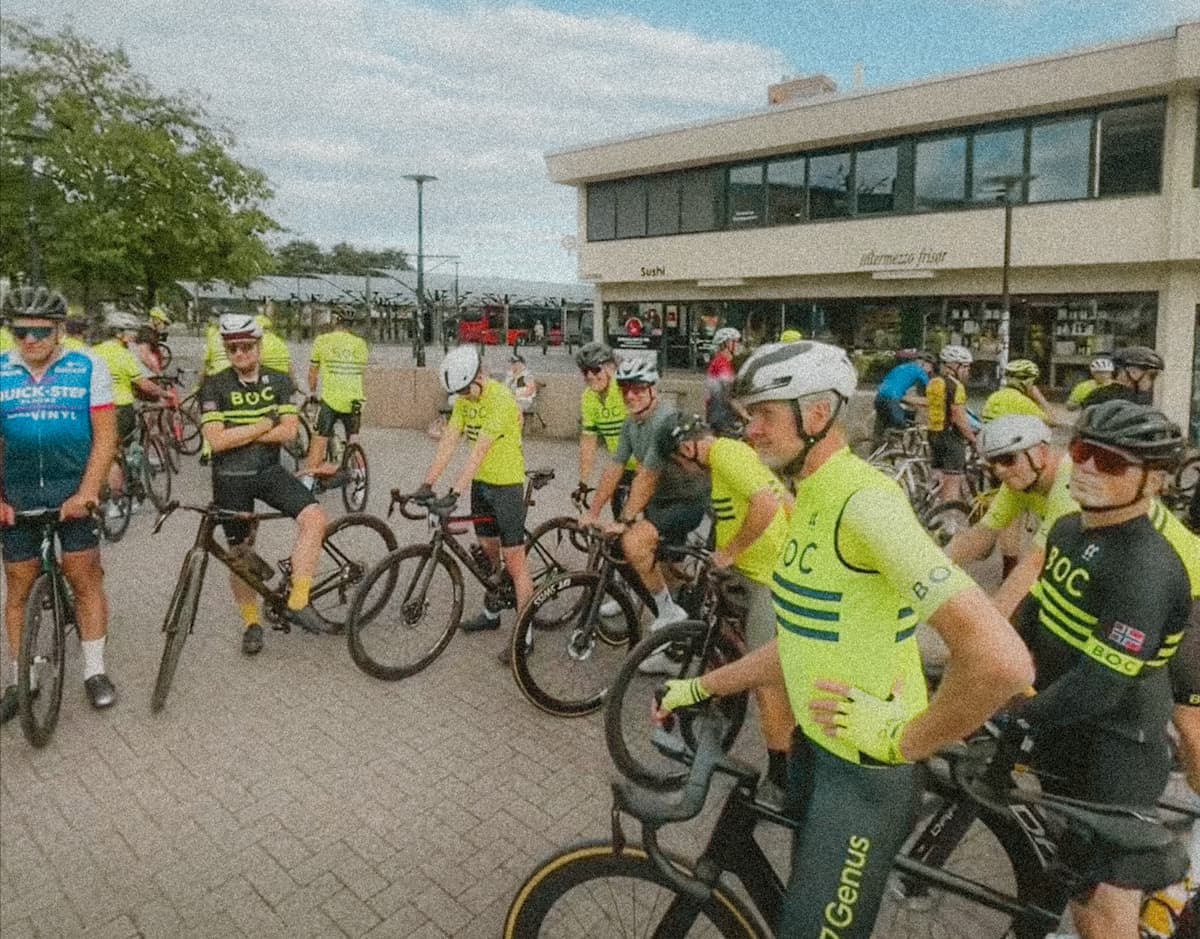 Ryttere i gul BOC-drakt står med syklene på Bekkestua torg før trening, med en kafébygning bak