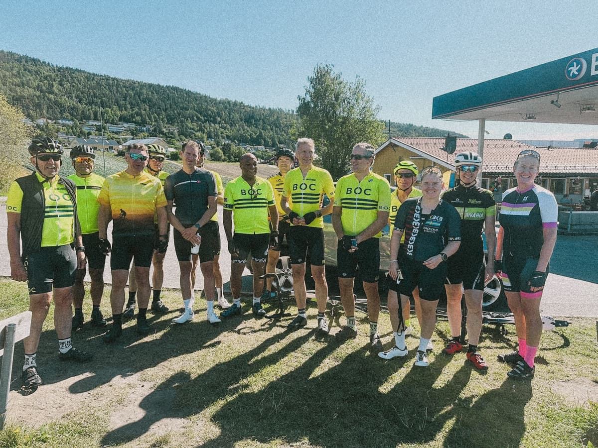 Ryttere i gul BOC-drakt og andre klubbdrakter på gresset ved en bensinstasjon, med åser og blå himmel bak