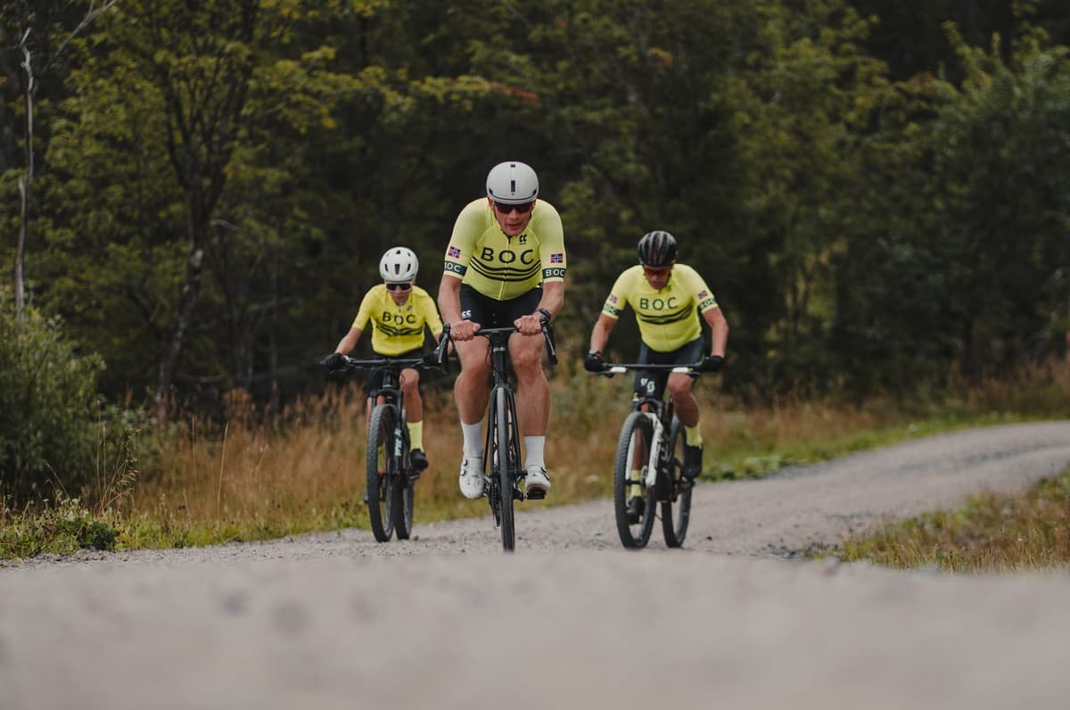 Tre syklister i gule drakter sykler sammen mot kameraet på grusveien