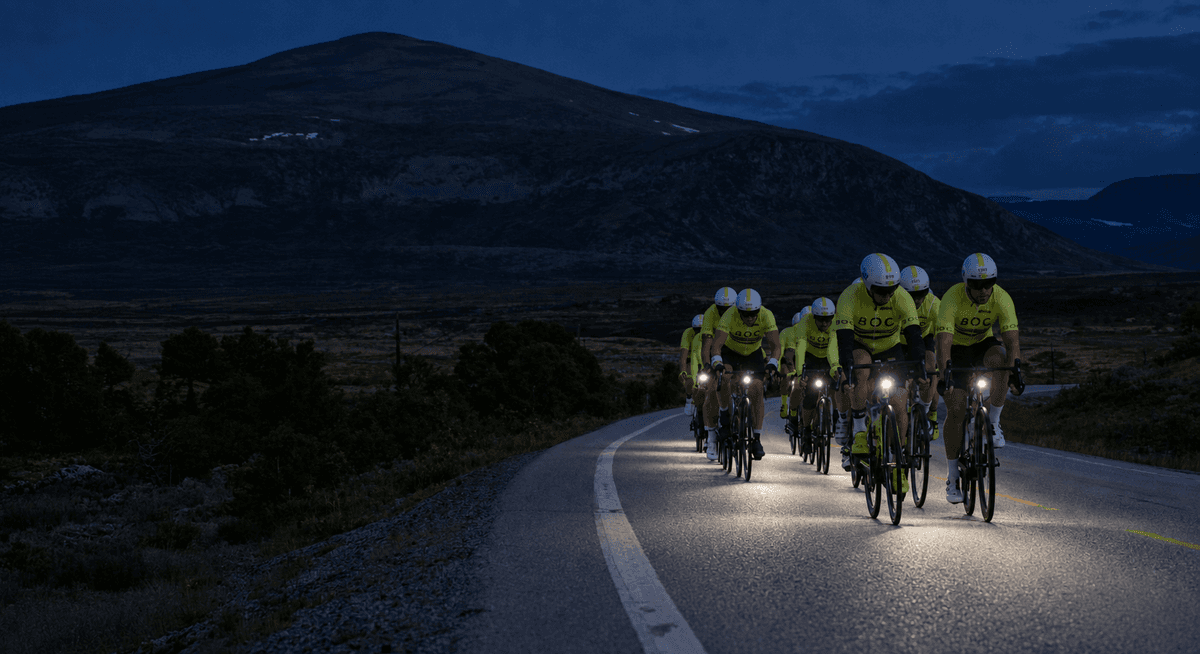 BOC-ryttere i gul klubbdrakt sykler samlet på en fjellvei i skumringen, med lys på syklene