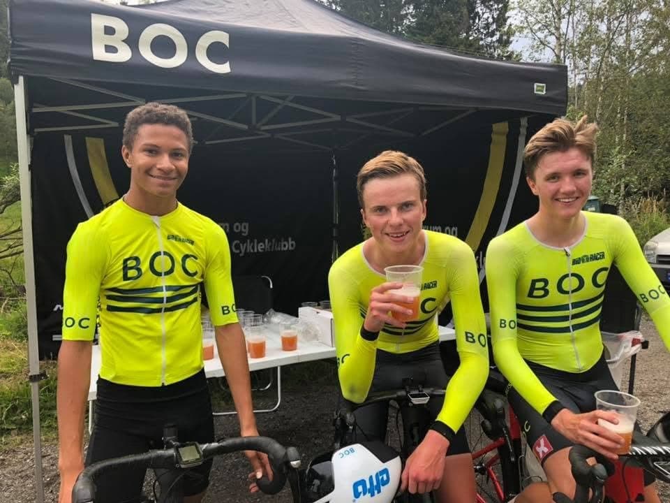 Tre unge ryttere i gul BOC-drakt smiler og drikker saft under BOC-teltet etter et ritt