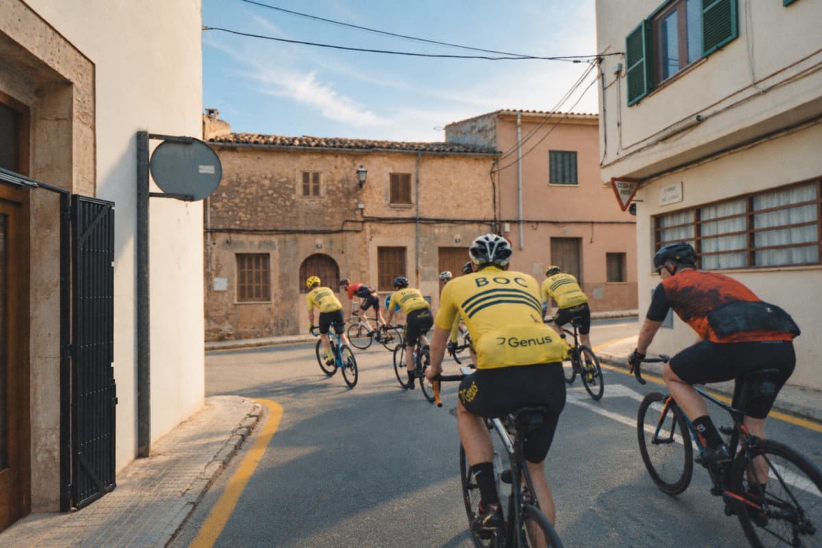 Syklister i gule BOC-drakter i en smal gate mellom gamle hus på Mallorca