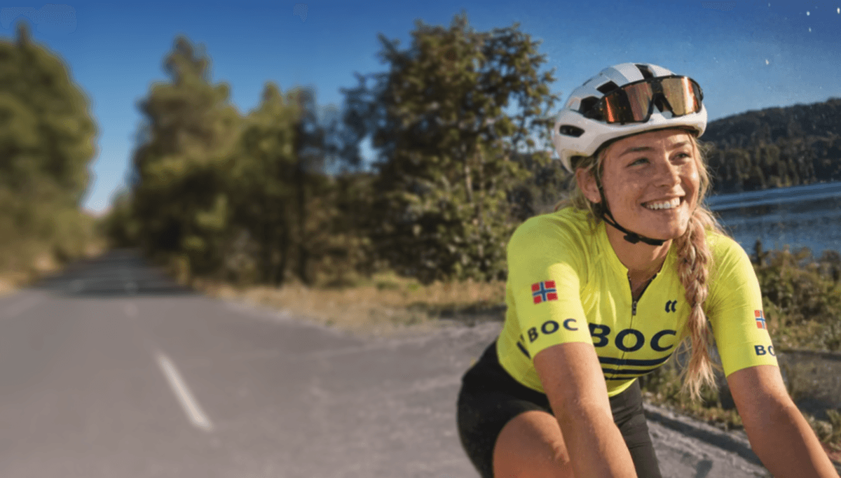 Smilende kvinne med hjelm, solbriller og gul BOC-drakt ved et vann