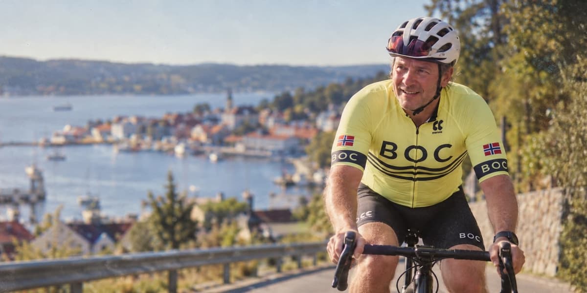 Smilende mann i gul BOC-drakt og hjelm på racersykkel, med en havneby i bakgrunnen